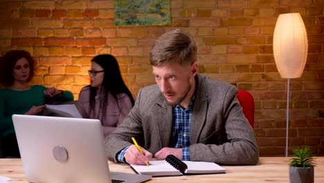 Closeup-shoot-of-adult-businessman-using-the-laptop-taking-notes-indoors-in-the-office.-Female-employee-using-the-tablet-sitting-on-the-couch-and-discussing-data-with-a-colleague-on-the-background