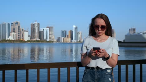 Hermosa-chica-con-el-pelo-largo-en-gafas-de-sol-usando-aplicación-de-teléfono-inteligente-en-Sunset-River-Quay-cerca-de-4K.