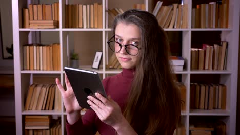 Nahaufnahme-Porträt-der-jungen-kaukasischen-Studentin-in-Brille,-die-auf-dem-Tablet-drinnen-in-der-College-Bibliothek-drinnen-tippt