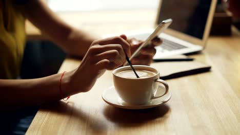 Primer-plano-de-las-manos-de-la-mujer-mezclando-café-en-taza-y-usando-teléfonos-inteligentes-relajándose-en-el-café