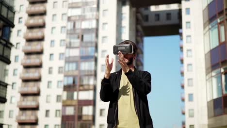 Tracking-slow-motion-shot-of-young-man-in-vr-glasses-standing-outdoors-in-city-street-and-exploring-virtual-reality