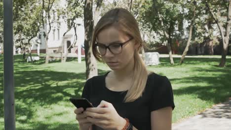 Perplexed-caucasian-woman-in-eyeglasses-walking-checking-smart-phone-content-and-then-stops-looking-at-camera-in-a-park