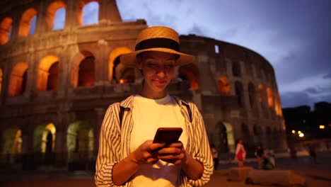 Alegre-turista-femenino-en-sombrero-satisfecho-con-conexión-4G-en-itinerancia-compartir-multimedia-y-charlar-en-línea-mientras-hace-turismo-en-Roma-cerca-del-coliseo-por-la-noche