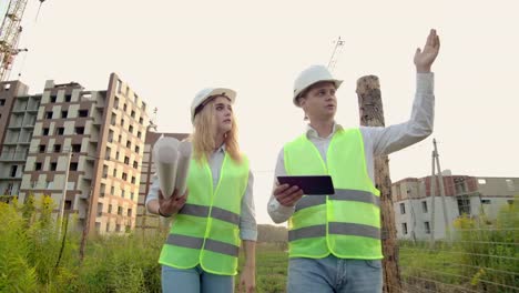 Negocios,-construcción,-industria,-tecnología-y-concepto-de-personas---constructor-sonriente-en-hardhat-con-tableta-PC-ordenador-junto-con-la-mujer-con-dibujos-de-constructores-en-el-sitio-de-construcción.