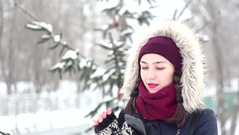 Hermosa-joven-está-volteando-a-través-de-las-redes-sociales-en-su-teléfono-inteligente-mientras-camina-en-un-parque-de-invierno.