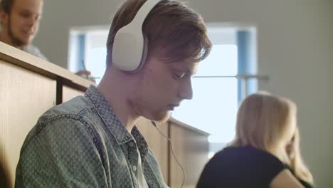 A-male-student-in-a-University-auditorium-listens-to-music-in-white-headphones-during-a-break-without-paying-attention-to-others.-A-lot-of-people-in-the-audience-are-talking