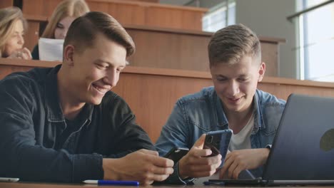 Männliche-Studenten-mit-einem-Smartphone-in-der-Hand-lachen-im-Publikum-während-einer-Pause-für-eine-Vorlesung-an-der-Universität