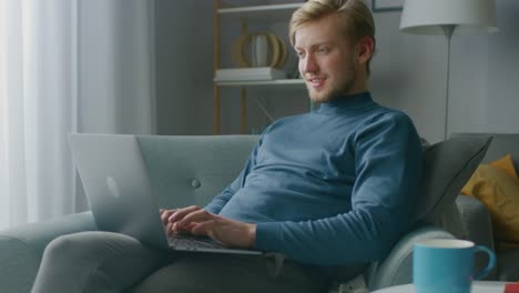 Porträt-von-Handsome-Blonde-Young-Man-Arbeitet-an-einem-Laptop-Computer,-während-auf-einem-Stuhl-in-seinem-gemütlichen-Wohnzimmer-sitzen.-Creative-Freelancer-entspannt-zu-Hause,-surft-im-Internet,-nutzt-Social-Media-und-entspannt