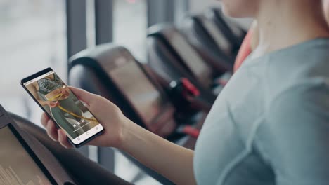 Primer-plano-de-una-mujer-usando-Smartphone-en-un-gimnasio-en-una-cinta-de-correr,-navegando-a-través-de-videos-motivacionales-en-la-red-social.-Tocar-con-el-dedo-entre-vídeos-en-la-fuente-de-aplicaciones-sociales.-Diseño-de-aplicaciones-simulada.