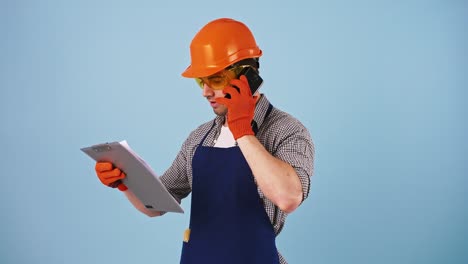 Young-man-in-safety-clothes,-hard-hat,-protective-goggles,-gloves.-Talking-by-smartphone,-looking-at-papers-on-clipboard.-Posing-on-blue-background