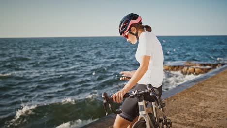 Girl-in-protective-helmet,-sunglasses-and-sportswear-is-sitting-on-bike-at-sea-embankment.-She-is-typing-on-her-cellphone.-Sunny-day,-slow-motion