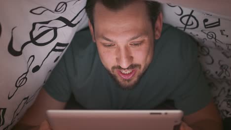 Close-up-of-face-of-a-young-bearded-and-mustashed-man,-using-digital-tablet-under-blanket,-who-is-chatting-and-waves-hand,-in-his-bed,-at-night