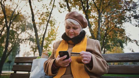 Rentnerin-betritt-eine-Reihe-ihrer-Kreditkarte-ins-Handy,-während-sie-im-Herbstpark-auf-der-Bank-sitzt