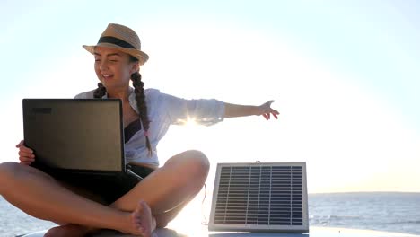 tourism,-girl-sits-on-car-roof-in-backlight-speaks-on-laptop-and-recharges-solar-array,-young-woman-sitting-on-vintage-car