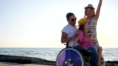 happy-invalid,-family-play-together-on-background-blue-sky,-girl-sitting-on-daddy-in-wheelchair-with-outstretched-arms