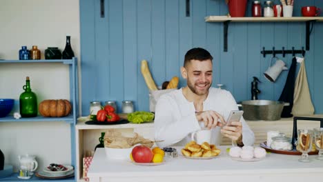 Fröhlicher-junge-Mensch-mit-Smartphone-in-der-Küche-sitzen-während-haben-das-Frühstück-am-Morgen