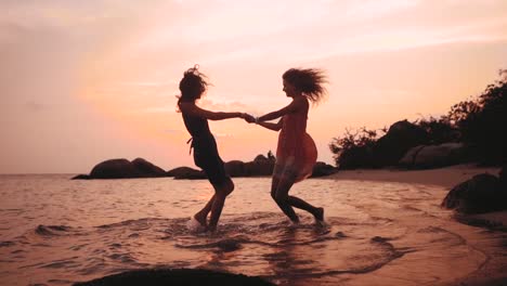 zwei-glückliche-tanzende-Mädchen-Hand-in-Hand-am-Strand-bei-Sonnenuntergang-in-Zeitlupe