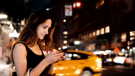 Portrait-of-young-happy-woman-standing-in-traffic-downtown-in-the-evening-in-New-York,-America-and-using-the-smartphone