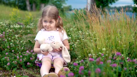 A-little-girl-playing-with-a-toy-rabbit-in-the-meadow-among-the-flowering-clover