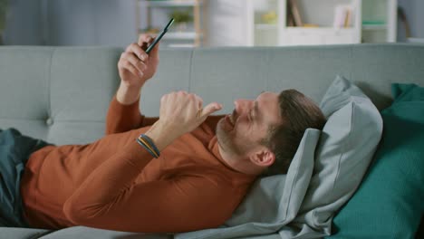 Mann-zu-Hause-liegen-auf-Sofa-macht-Videokonferenz-am-Smartphone.