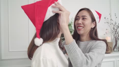 Pareja-de-lesbianas-asiáticas-dar-y-usar-sombrero-de-la-Navidad-uno-al-otro-en-su-sala-de-estar-en-el-país-en-el-Festival-de-Navidad.-Las-mujeres-del-estilo-de-vida-lgbt-felizes-celebran-Navidad-y-año-nuevo-concepto.