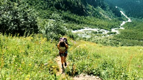 Young-woman-tourist-with-a-backpack-rises-uphill-in-the-background-of-a-beautiful-landscape.-The-girl-climbs-in-the-summer-on-foot