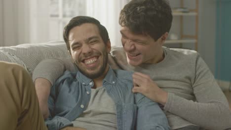 Portrait-of-a-Cute-Male-Queer-Couple-at-Home.-They-Sit-on-a-Sofa-and-Look-at-the-Camera.-Partner-Embraces-His-Lover-from-Behind.-They-are-Happy-and-Smiling.-Room-Has-Modern-Interior.