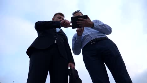 Two-young-businessmen-talking-and-using-tablet-pc-in-city.-Handsome-coworkers-discussing-and-working-on-digital-tablet-with-sky-at-background.-Colleagues-applying-mobile-technology.-Low-angle-view