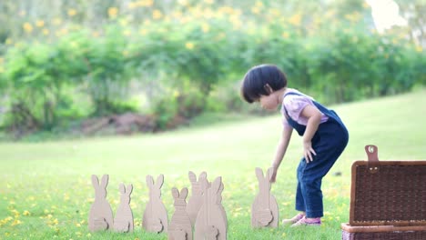 Niedliches-kleines-Kindermädchen-am-Ostersamstag.-Mädchen-jagt-nach-Ostereiern-auf-dem-Rasen-und-Hase-Made-of-paper-in-der-Natur-oder-Park-und-Sonnenlicht.-Zeitlupe