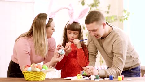 mamá,-papá-e-hija-apoyándose-en-la-mesa-decoran-los-huevos-de-Pascua.