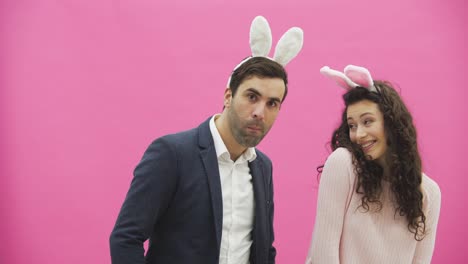 Beautiful-young-couple-standing-on-pink-background.-At-the-same-time,-the-girl-claps-her-husband,-he-in-turn-bites-her-hand.-Together-with-pink-barking-ears-on-the-head.-Happy-family-is-preparing-for-Easter.