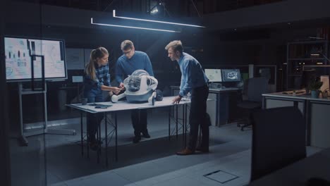 Tarde-en-la-noche-en-el-centro-de-ingeniería-de-robótica-tres-ingenieros-técnicos-hablan-y-trabajan-en-un-prototipo-de-robot-con-ruedas.-En-el-fondo-centro-de-investigación-de-alta-tecnología-con-pantallas-que-muestran-diseño-industrial