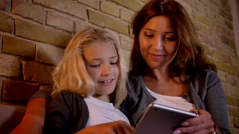 Closeup-shoot-of-young-caucasian-mother-and-her-small-pretty-girl-using-the-tablet-together-while-sitting-on-the-couch-indoors-at-home