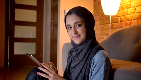 Closeup-shoot-of-young-attractive-muslim-female-in-hijab-typing-on-the-phone-while-sitting-on-the-floor-in-the-doorway-at-cozy-home