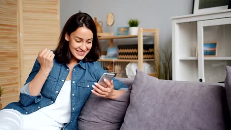 Jovencita-emocionada-celebrando-el-éxito-mirando-a-la-pantalla-del-teléfono-inteligente-en-casa