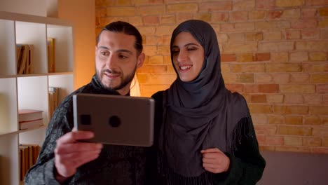 Closeup-shoot-of-young-cheerful-muslim-couple-having-a-video-call-on-the-tablet-and-showing-off-their-newly-bought-apartment