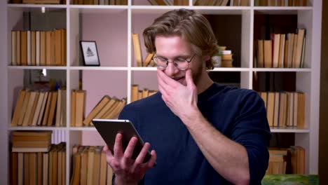 Closeup-shoot-of-adult-attractive-male-student-using-the-tablet-smiling-happily-in-the-university-library-indoors