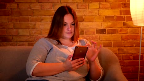 Porträt-von-entspannten-Plus-Size-Modell-Fixierung-ihre-Haare-halten-ein-Smartphone-in-gemütlicher-Häuslicheatmosphäre.