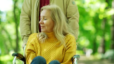 Zoom-in-of-male-caregiver-or-husband-pushing-senior-woman-in-wheelchair-towards-camera-in-park.-Sad-disabled-woman-looking-at-camera