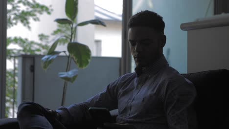 Young-afro-american-Businessman-taking-his-smartphone-in-his-living-room.-Social-network-account-feed,-news.-Medium-Shot.-Communication,-social-networking,-online-shopping,-chat-concept.