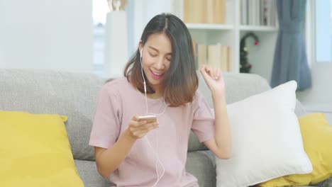 Asian-woman-listening-music-and-using-smart-phone,-female-using-relax-time-lying-on-home-sofa-in-living-room-at-home.-Happy-female-listening-music-with-headphones-concept.