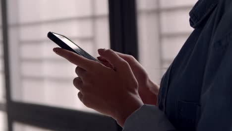 Close-up-hands-young-asian-business-woman-holding-typing-mobile-phone-and-scrolls-through-social-media-feed-in-smartphone-standing-beside-window-at-home-office.