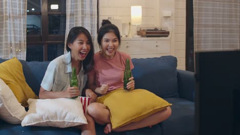 Lesbian-lgbt-women-couple-party-at-home,-Asian-female-drinking-beer-watching-TV-cheer-soccer-funny-moment-together-on-sofa-in-living-room-in-night.-Young-lover-football-fan,-celebrate-holiday-concept.