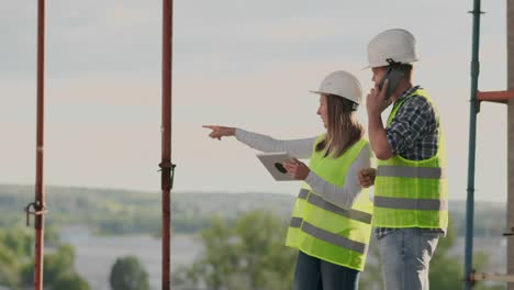 Ingenieure-Designer-stehen-auf-dem-Dach-des-Gebäudes-im-Bau-und-diskutieren-den-Plan-und-den-Fortschritt-des-Baus-mit-einem-Tablet-und-Handy