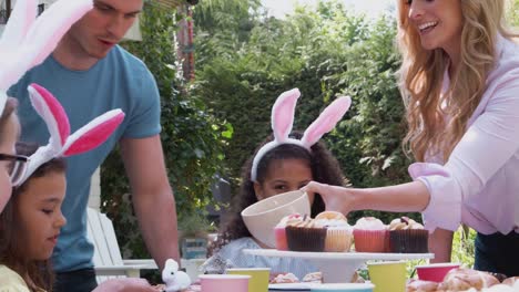 Grupo-de-niños-con-orejas-de-conejo-sentados-en-la-mesa-al-aire-libre-disfrutando-de-la-fiesta-de-Pascua-con-los-padres---tiro-a-cámara-lenta