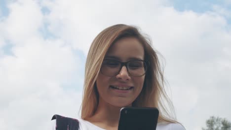 Una-mujer-caucásica-sonriente-con-gafas-camina-por-el-parque-de-la-ciudad-y-usa-su-teléfono.-Bonita-mujer-de-verano-en-blanco-t-corto-camina-por-la-calle-mirando-a-su-teléfono-móvil