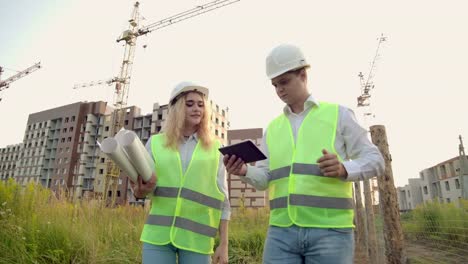 Ein-Mann-und-eine-Frau-in-Schutzkleidung-und-Helmen-gehen-auf-die-Baustelle,-um-zu-diskutieren-und-sich-umzusehen-klicken-Klicken-Sie-auf-dem-Tablet-Bildschirm-auf-dem-Hintergrund-der-Arbeitskrane