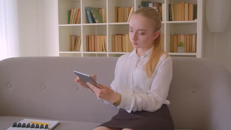 Retrato-de-primer-plano-de-joven-bonita-estudiante-rubia-caucásica-usando-la-tableta-sentada-en-el-sofá-interior-en-el-apartamento