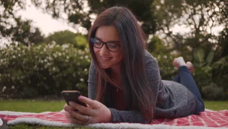 Retrato-de-joven-sonriente-relajada-y-tendida-en-la-manta-en-el-parque-usando-el-teléfono-inteligente