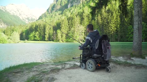 4k-Auflösung-von-behinderten-Fischer-in-einem-elektrischen-Rollstuhl-Angeln-in-schönen-See-in-der-Nähe-von-Wald-und-Berg-im-Rücken